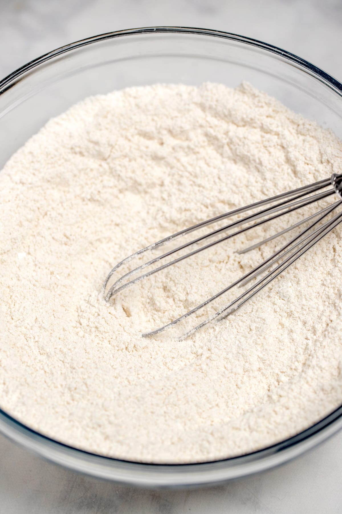 A whisk mixing together flour and other dry ingredients in a large glass mixing bowl on a table.