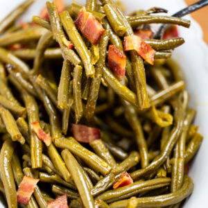 A bowl of crockpot green beans on a table, with a spoon lifting up a scoop of green beans topped with bacon.