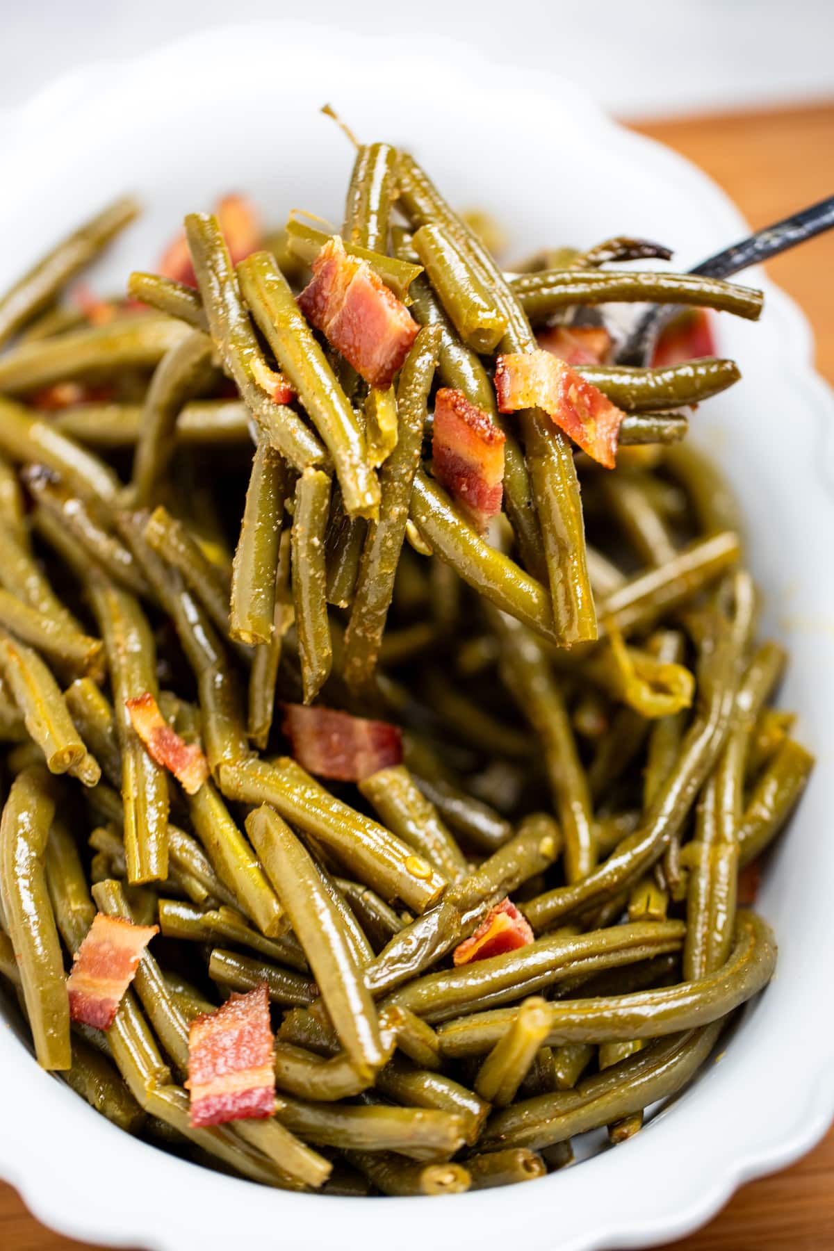 A bowl of crockpot green beans on a table, with a spoon lifting up a scoop of green beans topped with bacon.