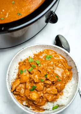 A bowl of slow cooker chicken tikka masala over rice, topped with fresh cilantro pieces, with a spoon on the table, in front of the slow cooker.