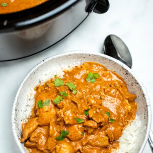 A bowl of slow cooker chicken tikka masala over rice, topped with fresh cilantro pieces, with a spoon on the table, in front of the slow cooker.