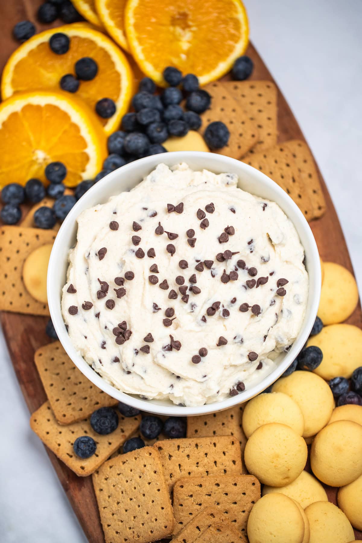 A bowl of cannoli dip topped with mini chocolate chips on a cutting board surrounded by graham crackers, vanilla wafers, blueberries, and orange slices.