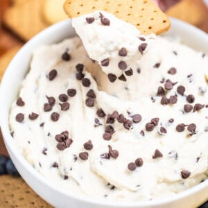A bowl of cannoli dip topped with mini chocolate chips, with a hand using a graham cracker to scoop up some dip. The bowl is sitting on a cutting board surrounded by graham crackers, blueberries, and vanilla wafers.