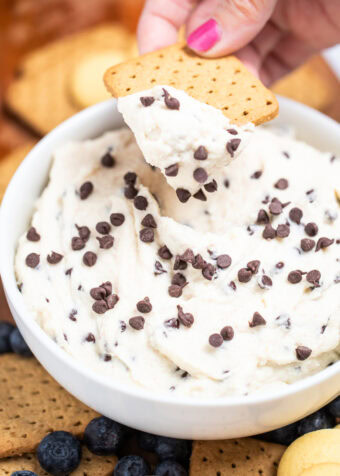 A bowl of cannoli dip topped with mini chocolate chips, with a hand using a graham cracker to scoop up some dip. The bowl is sitting on a cutting board surrounded by graham crackers, blueberries, and vanilla wafers.