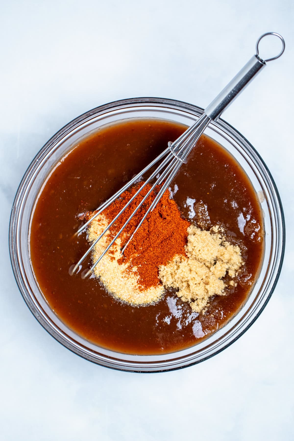 A glass bowl on a table with BBQ sauce, spices, and brown sugar waiting to be mixed together with a whisk.