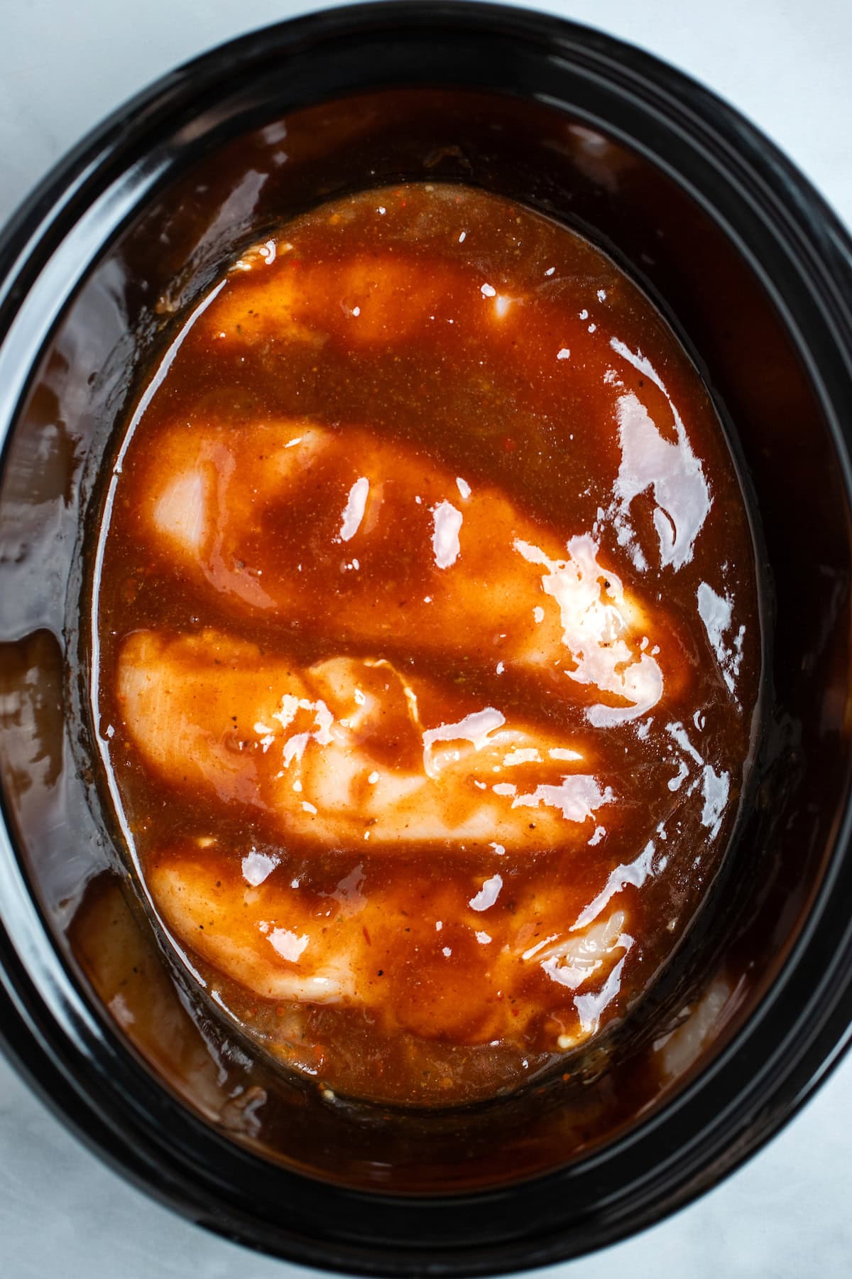 A slow cooker with raw chicken breasts side by side topped with BBQ sauce.