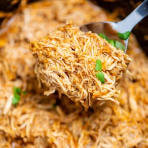 A slow cooker with saucy shredded BBQ chicken and a spoon lifting up the chicken.