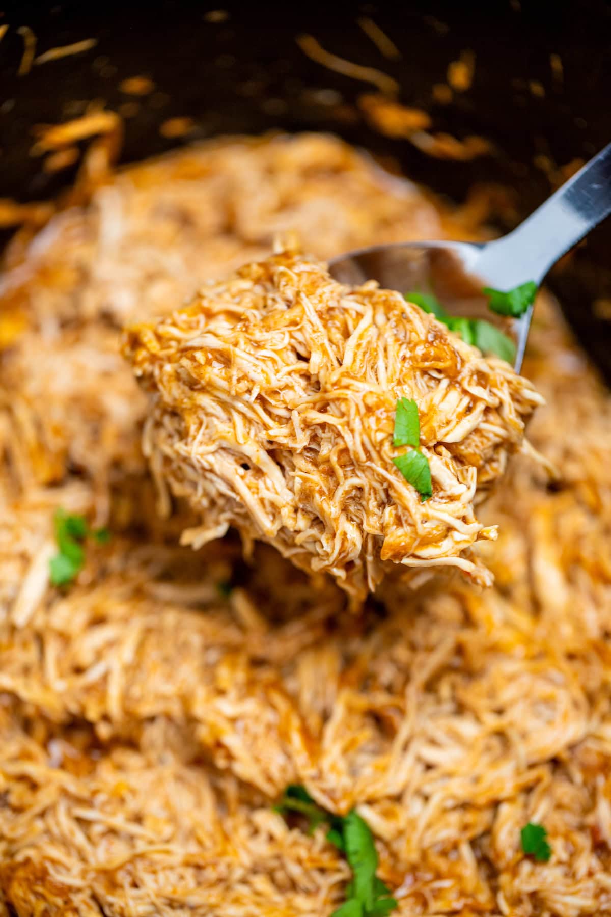 A slow cooker with saucy shredded BBQ chicken and a spoon lifting up the chicken.