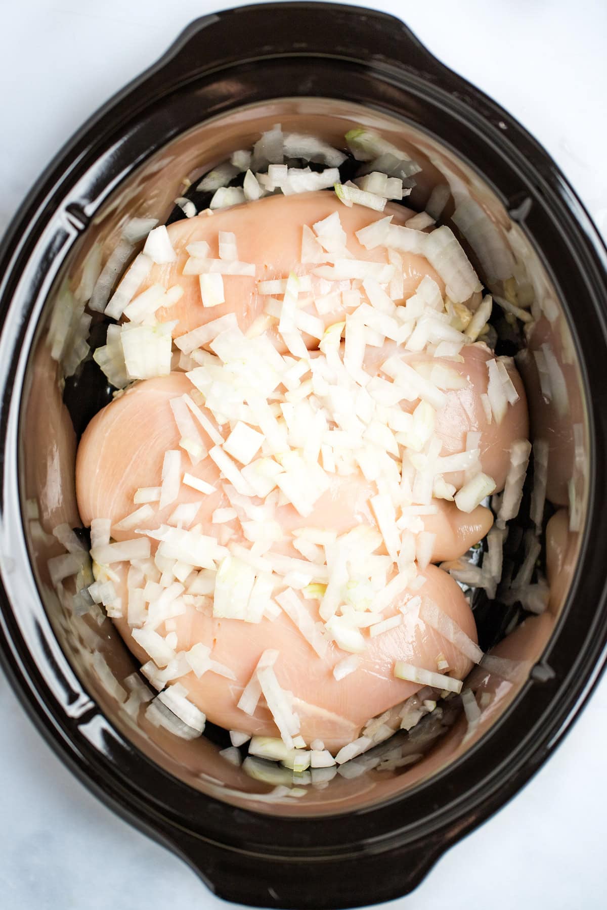 A slow cooker with raw chicken breast and chopped onion.