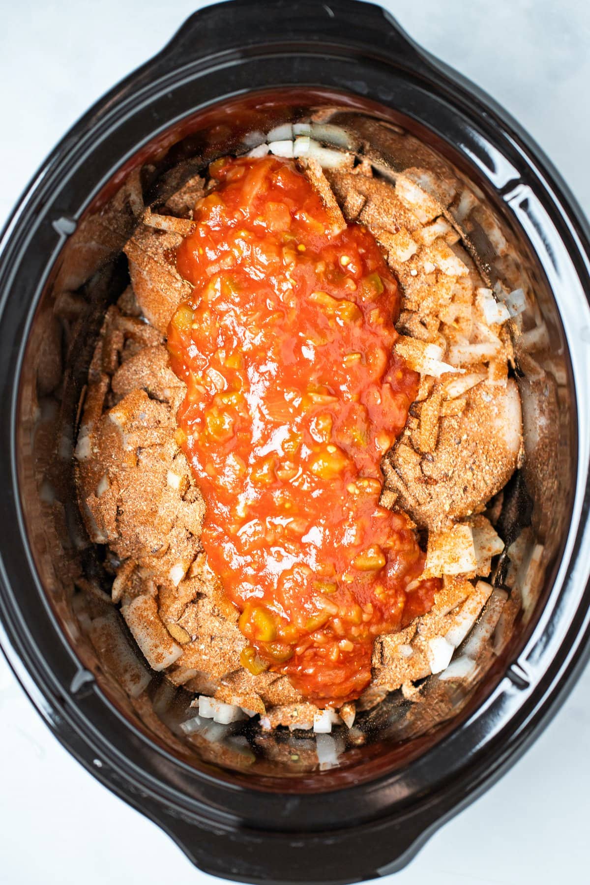 A slow cooker with raw chicken breast and chopped onion with taco seasoning and salsa on top.