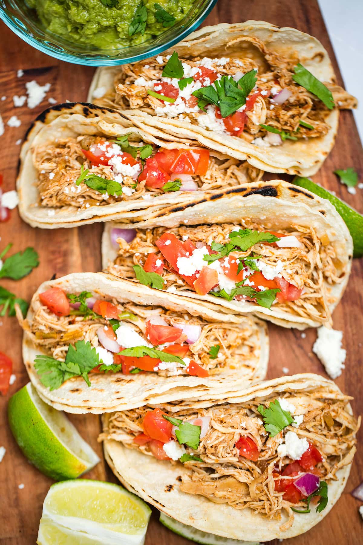 A cutting board with five slow cooker chicken tacos in corn tortillas topped with tomato, red onion, queso fresco, and cilantro, next to a bowl of guacamole and lime wedges.