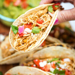 A hand holding a taco in a corn tortilla topped with tomato, red onion, queso fresco, and cilantro, above a cutting board with more tacos and a bowl of guacamole.