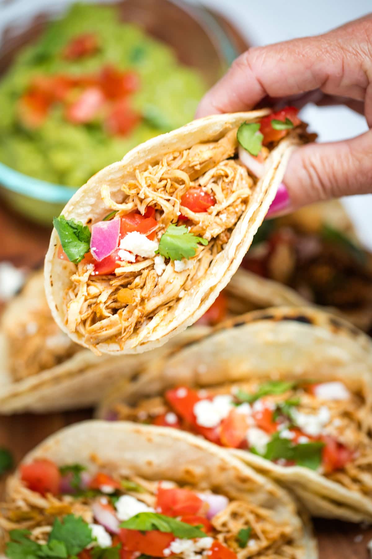 A hand holding a taco in a corn tortilla topped with tomato, red onion, queso fresco, and cilantro, above a cutting board with more tacos and a bowl of guacamole.