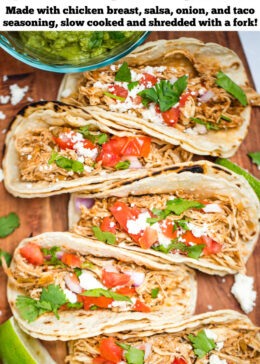 Pinterest pin with a cutting board with five slow cooker chicken tacos in corn tortillas topped with tomato, red onion, queso fresco, and cilantro, next to a bowl of guacamole and lime wedges.