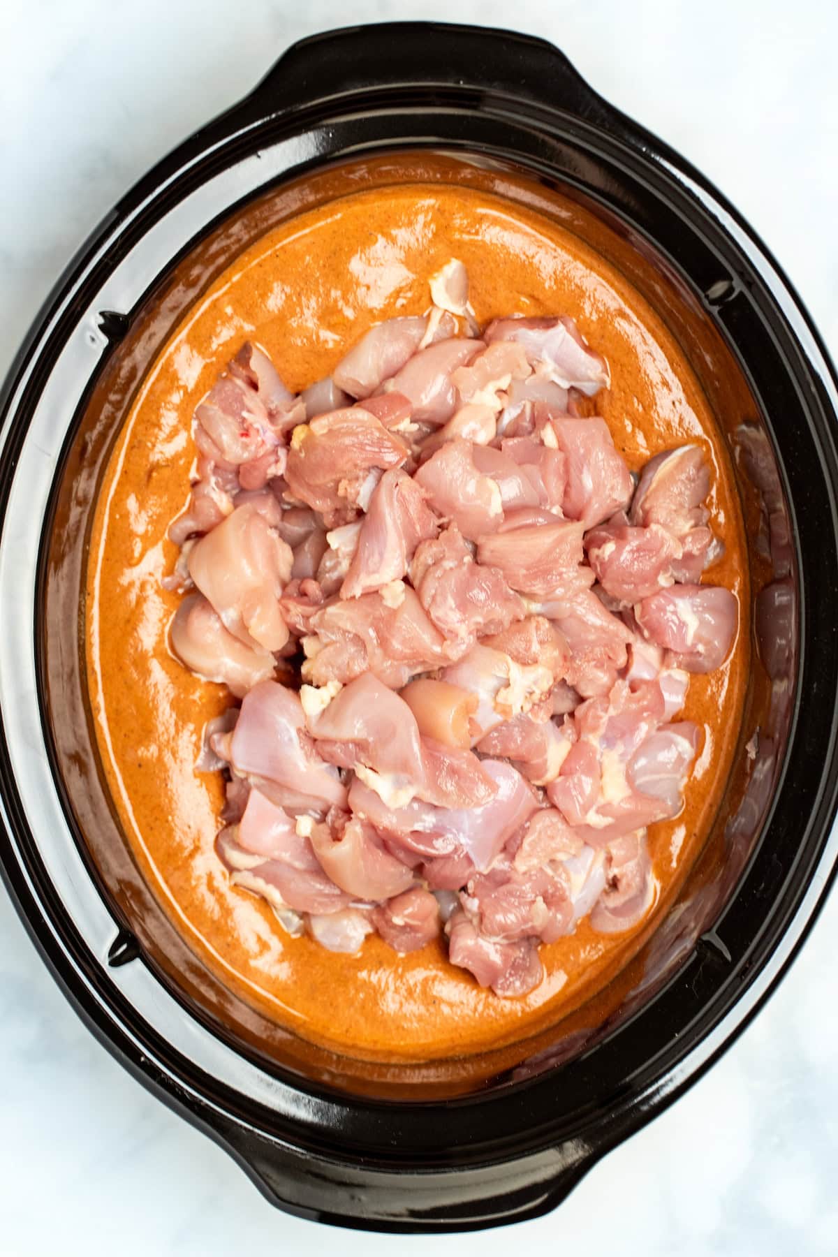 A slow cooker dish with tomato based sauce filling the bottom of the pot with raw pieces of chicken resting on top.
