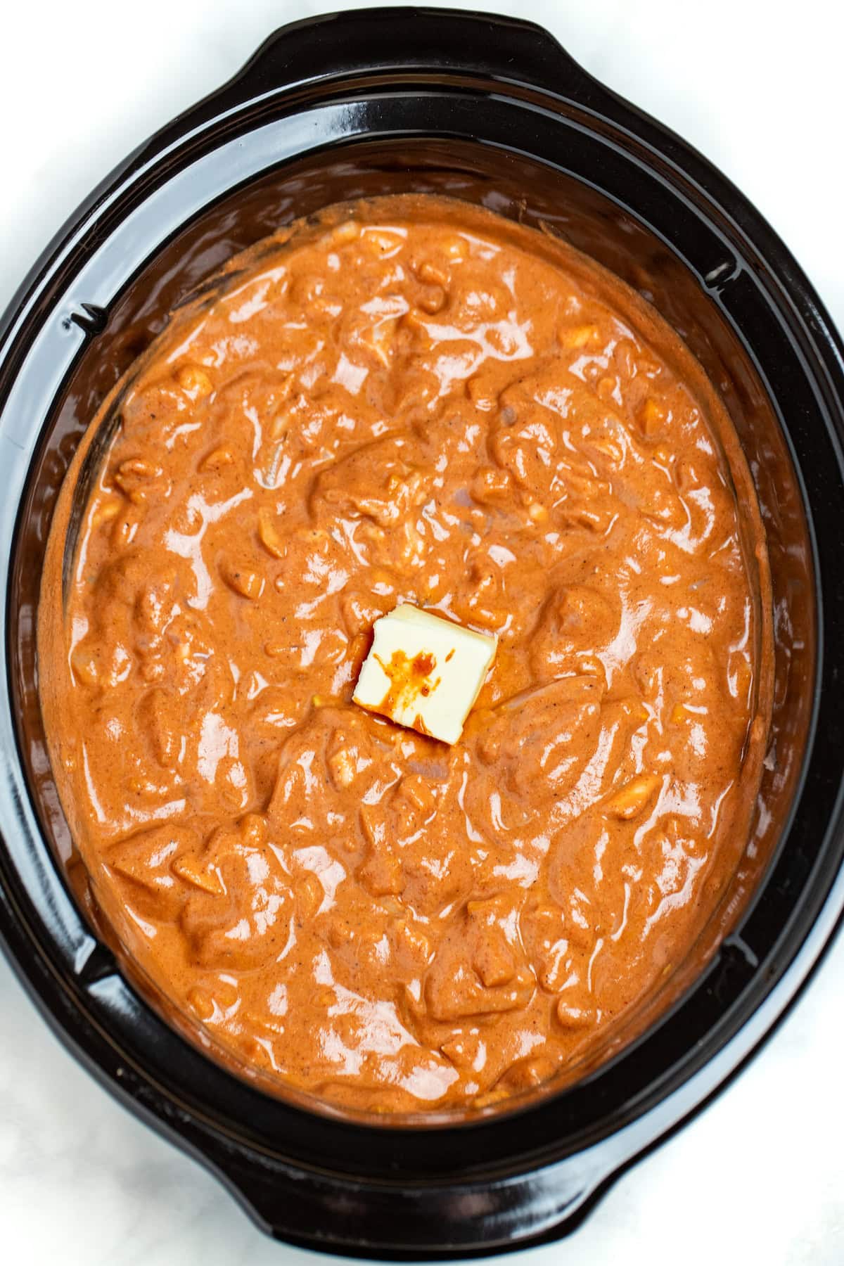 A slow cooker pot filled with uncooked chicken tikka masala, which is chicken, onion, bell pepper, and spices all mixed together, with a piece of butter resting on top.