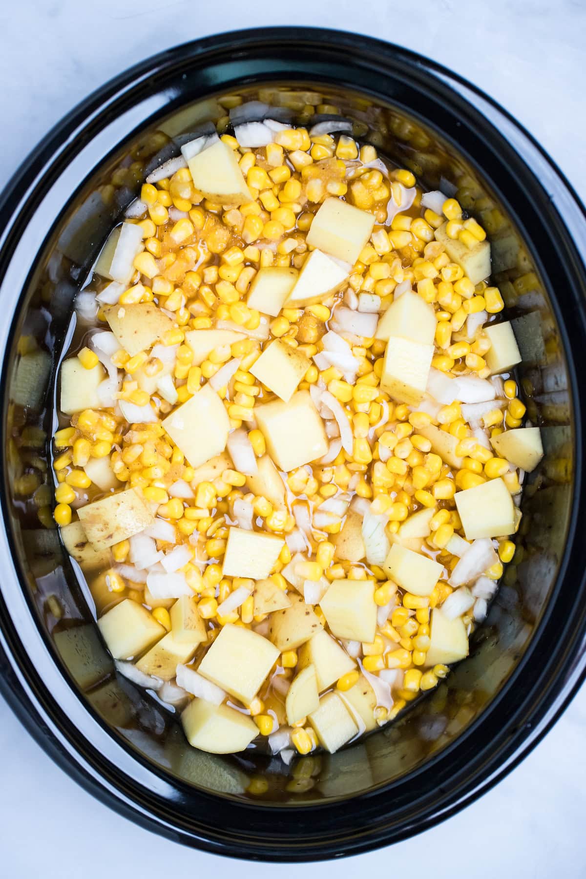 A slow cooker with broth, onion, corn kernels, and potatoes before cooking.