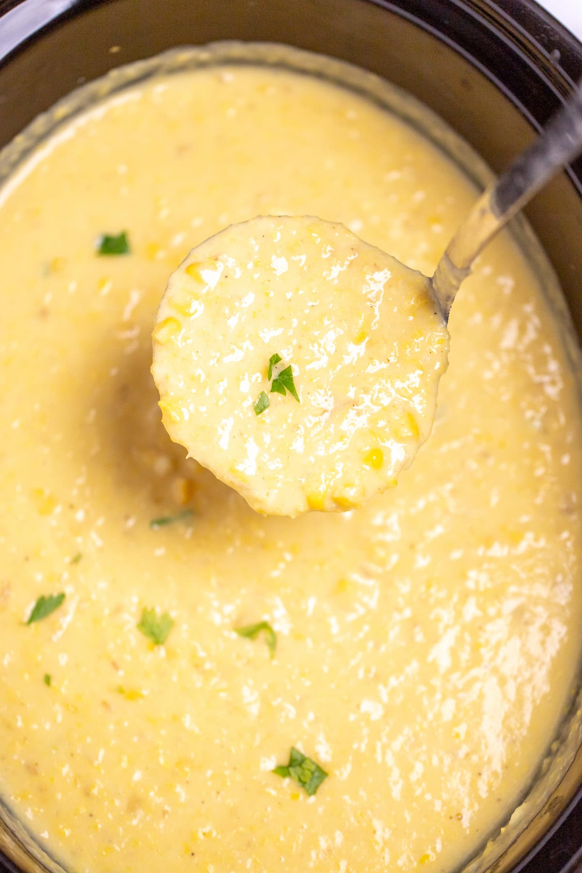 A slow cooker full of corn chowder topped with fresh chopped parsley, and a ladle scooping up some chowder.