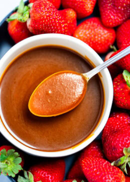 A spoon full of caramel sauce resting across a bowl of caramel sauce, with strawberries on a plate surrounding the bowl.