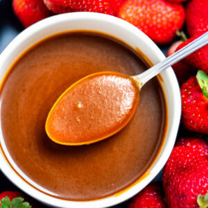 A spoon full of caramel sauce resting across a bowl of caramel sauce, with strawberries on a plate surrounding the bowl.