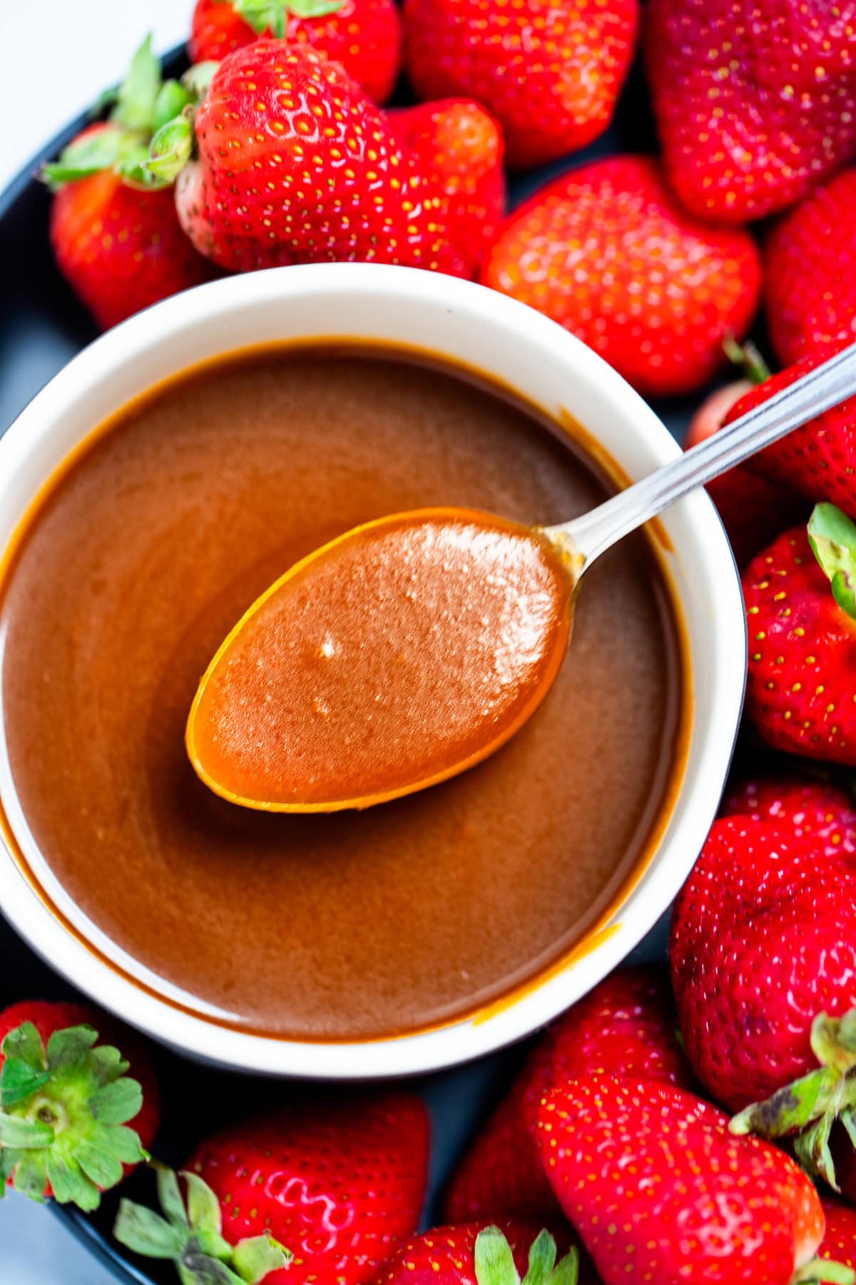 A spoon full of caramel sauce resting across a bowl of caramel sauce, with strawberries on a plate surrounding the bowl.