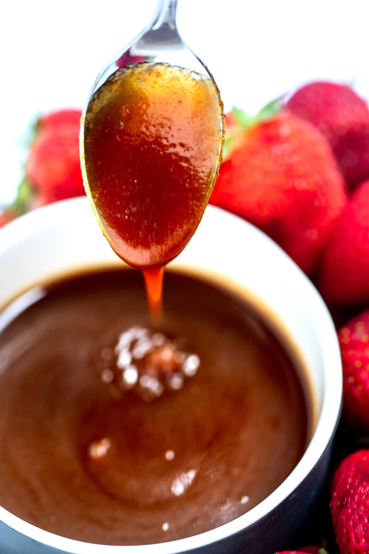 A spoon dipped into a small bowl of caramel sauce, being held above the dish with caramel dripping off the spoon back into the bowl. The bowl is surrounded by strawberries in the background.