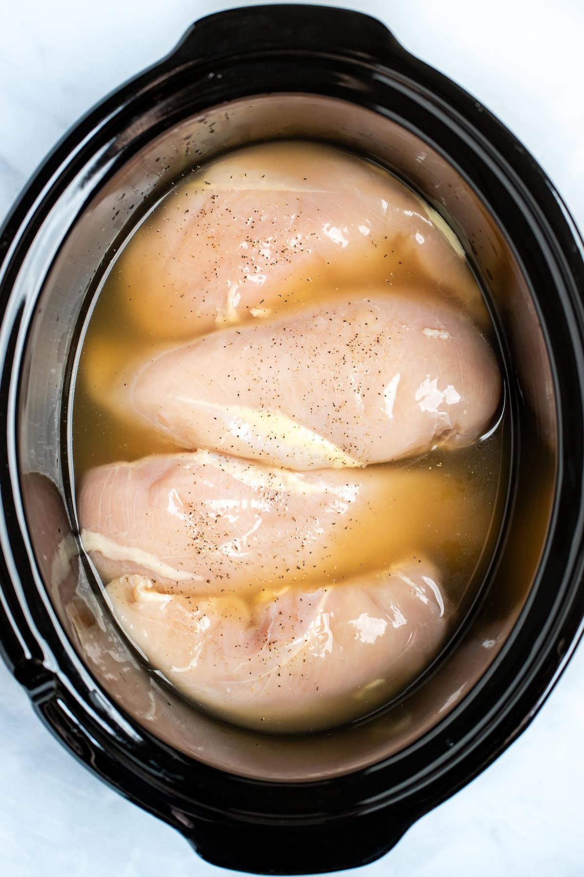 Medium sized raw chicken breasts in a slow cooker with broth, salt, and pepper.