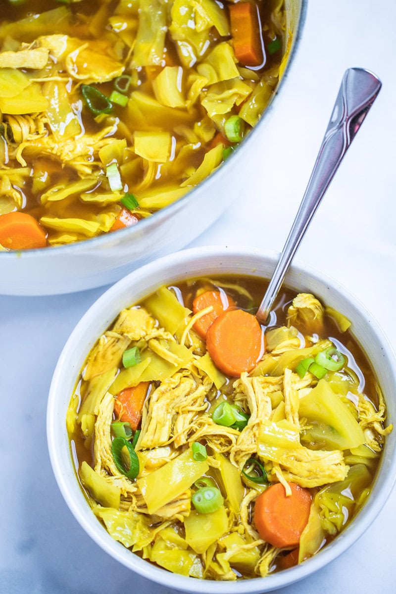 A bowl of cabbage chicken soup topped with green onions on a table with a spoon in the bowl, in front of the dutch oven of soup.