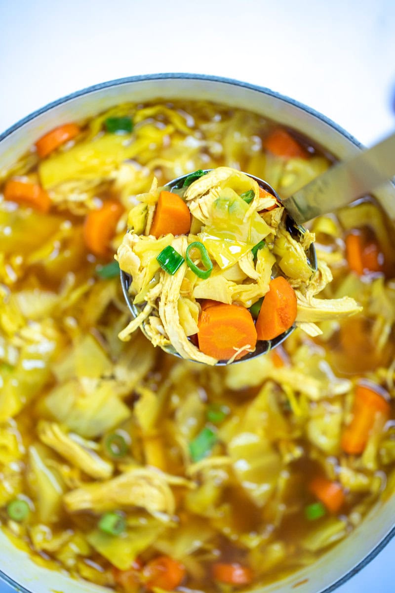 A dutch oven full of cabbage chicken soup topped with green onions, and a ladle scooping up the soup.