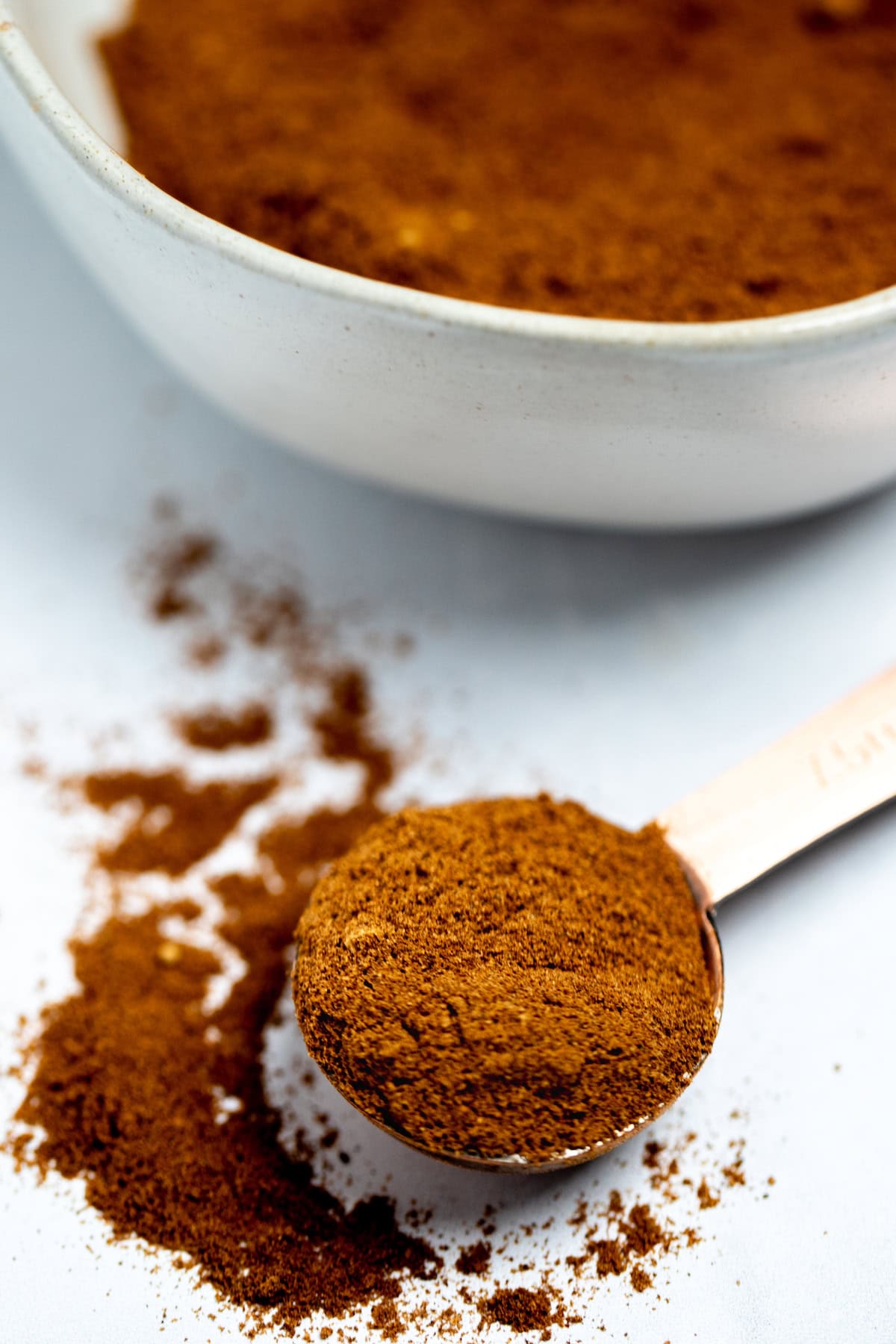 A tablespoon resting on a table, spilling pumpkin pie spice onto the table, with a small bowl in the background.