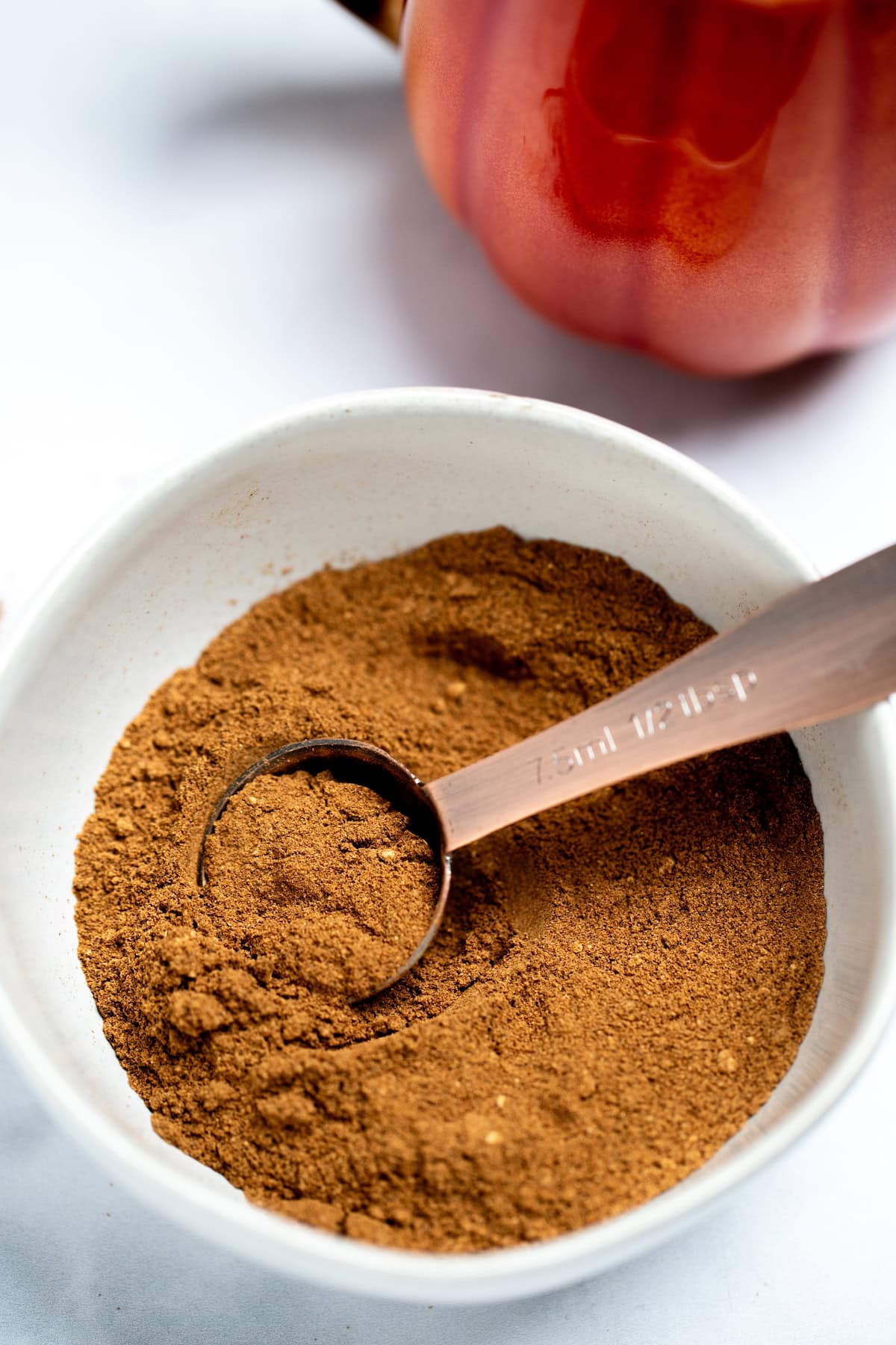 A small bowl with a tablespoon scooping some pumpkin pie spice up. There is a pumpkin shaped mug in the background.