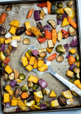 Roasted vegetables on top of parchment paper on a sheet pan, with a spatula in the middle lifting some vegetables up, just out of the oven.