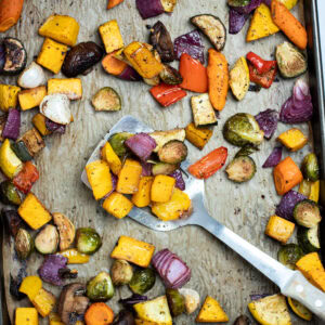 Roasted vegetables on top of parchment paper on a sheet pan, with a spatula in the middle lifting some vegetables up, just out of the oven.