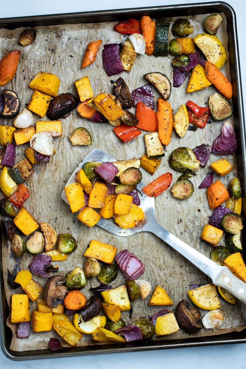 Roasted vegetables on top of parchment paper on a sheet pan, with a spatula in the middle lifting some vegetables up, just out of the oven.