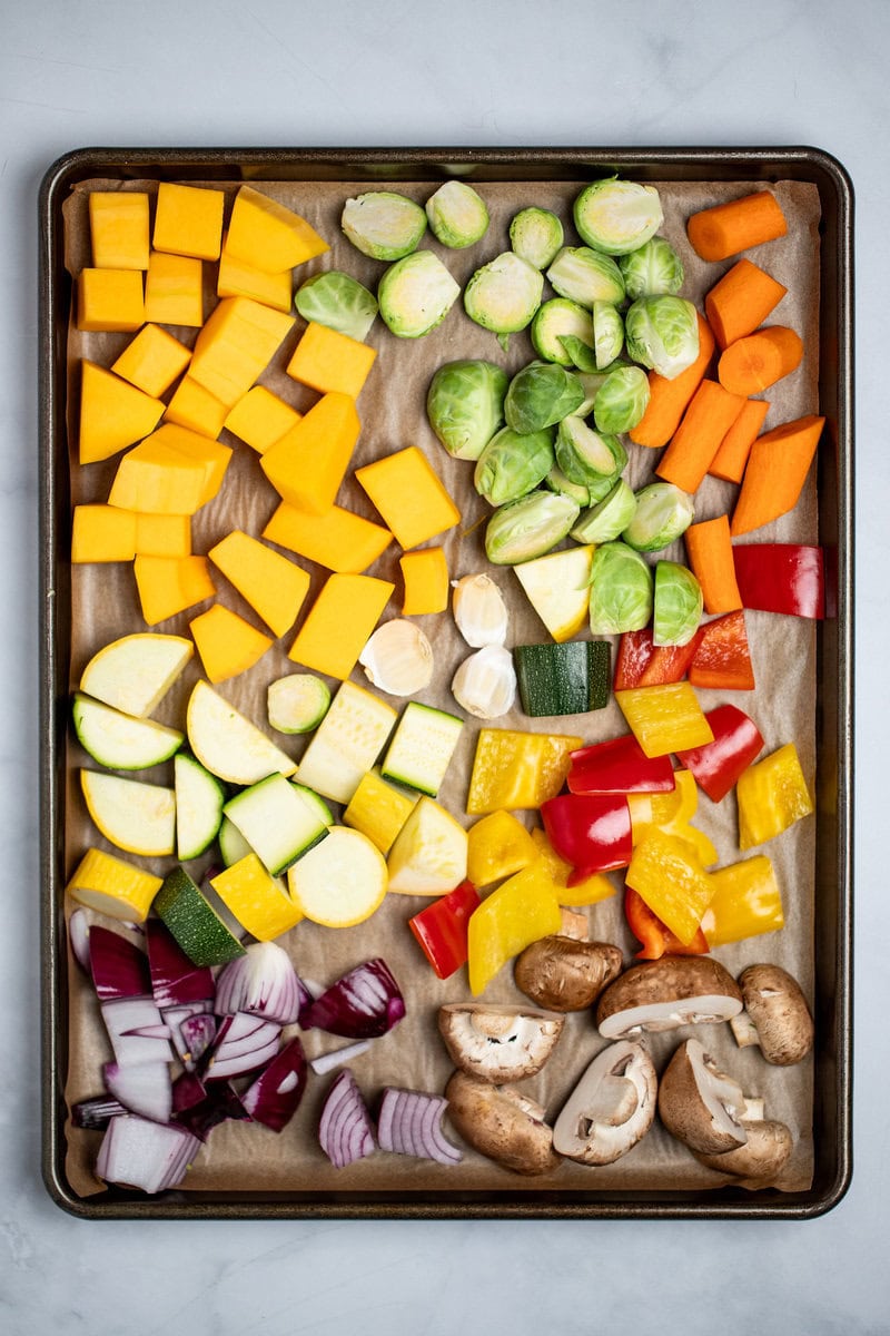 A sheet pan on a table with cut up mushrooms, onion, zucchini, yellow squash, bell pepper, Brussel sprouts, butternut squash, and carrots before adding the seasoning and roasting.