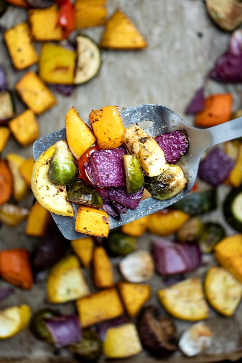 Roasted vegetables on top of parchment paper on a sheet pan, with a spatula in the middle lifting some vegetables up, just out of the oven.