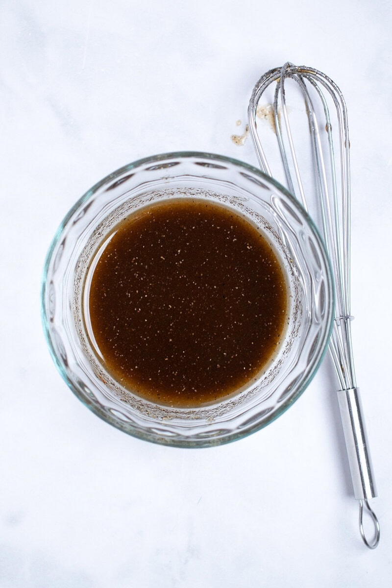 A small glass bowl with olive oil and spices whisked together, with a whisk laying next to the bowl.