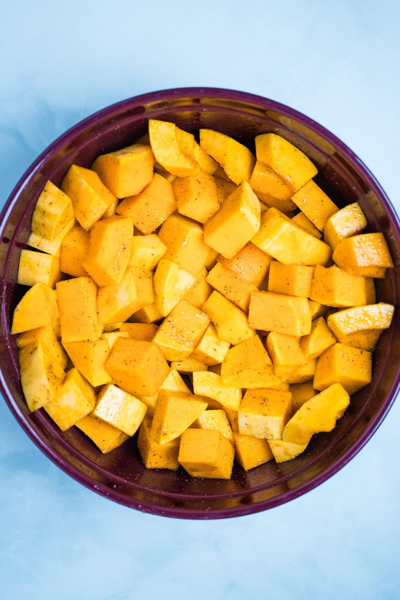 A bowl with cubed butternut squash covered in olive oil and spices, on a table.