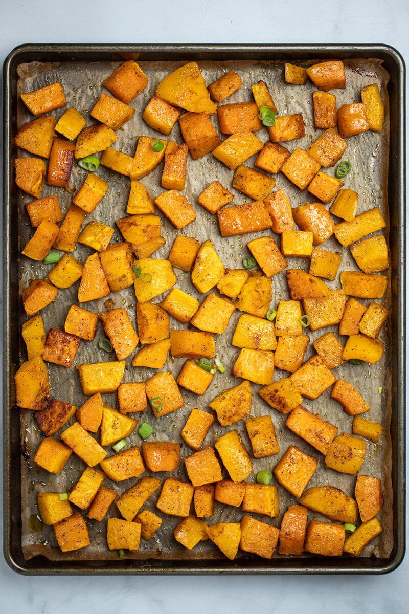 A sheet pan lined with parchment paper with a single layer of roasted butternut squash, topped with green onions.