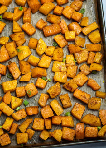 A sheet pan lined with parchment paper with a single layer of roasted butternut squash, topped with green onions.