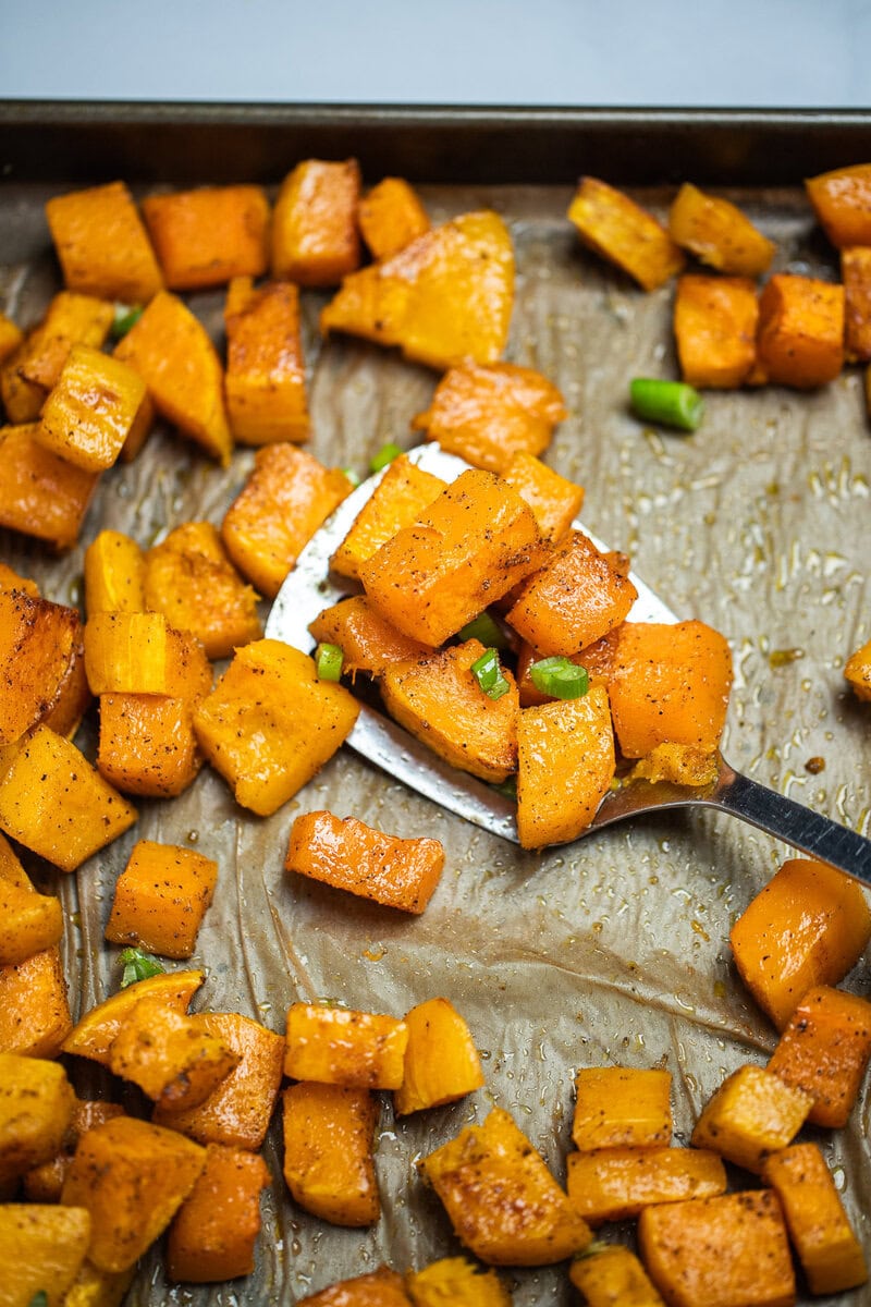 A sheet pan lined with parchment paper with a single layer of roasted butternut squash, topped with green onions, and a spatula lifting up some pieces of squash.