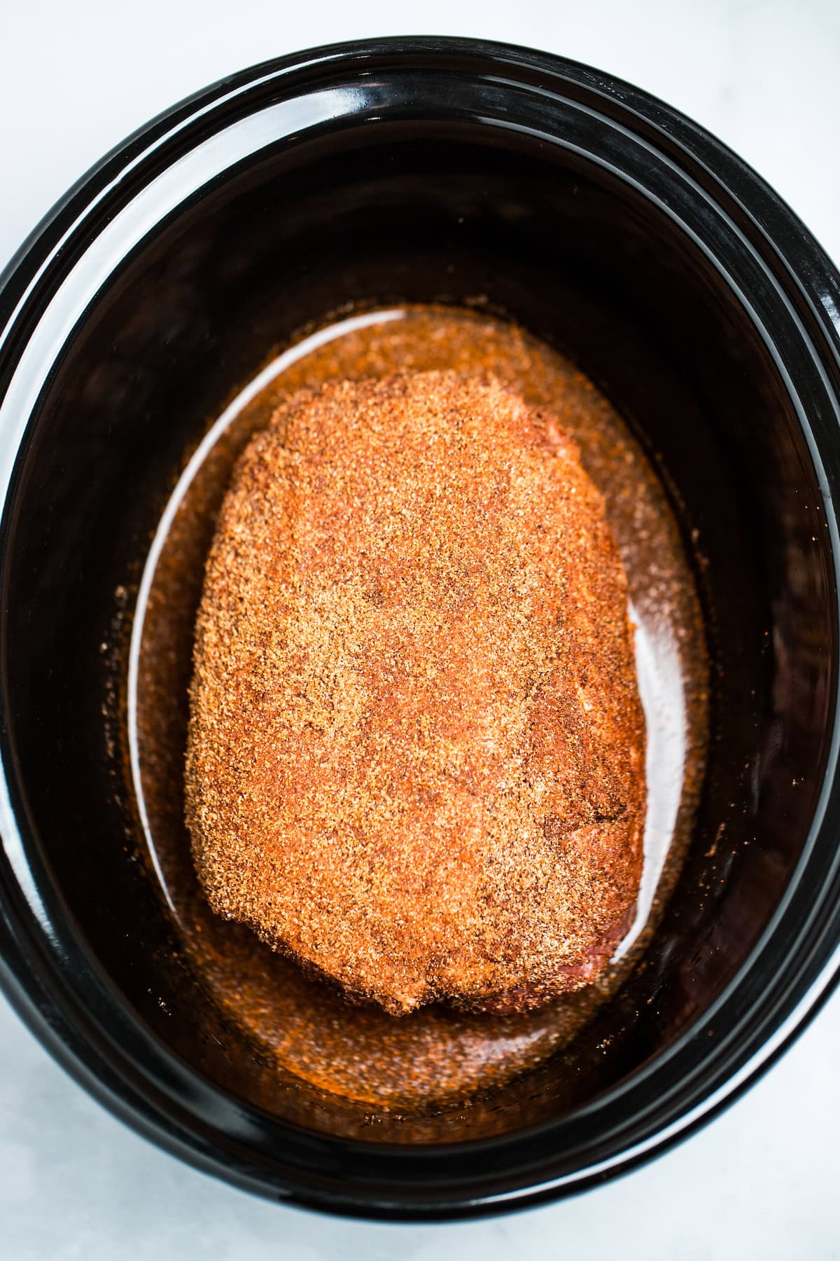 A slow cooker with broth and a pork butt rubbed with spices before cooking.