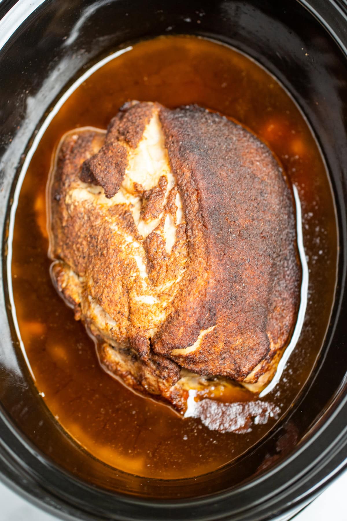 A slow cooker with broth and a pork butt rubbed with seasoning, fully cooked, before being shredded.