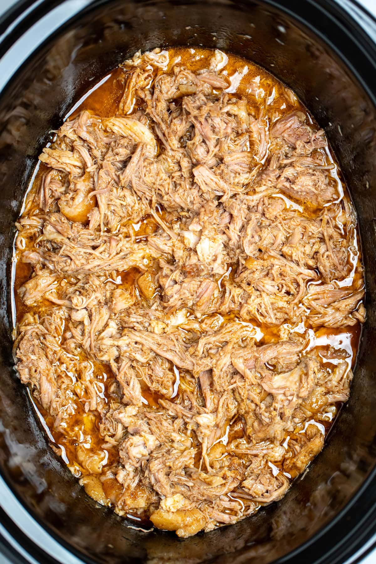 A slow cooker full of shredded pork shoulder in broth and drippings.