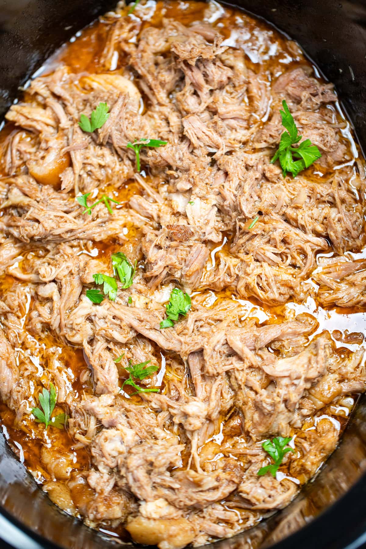 A slow cooker full of shredded pork shoulder in broth and drippings, topped with fresh parsley.