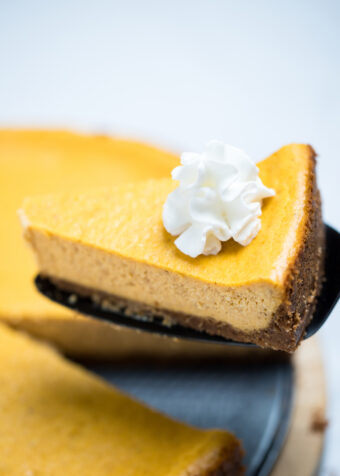 A piece of pumpkin cheesecake topped with whipped cream, being lifted up over the whole cheesecake.