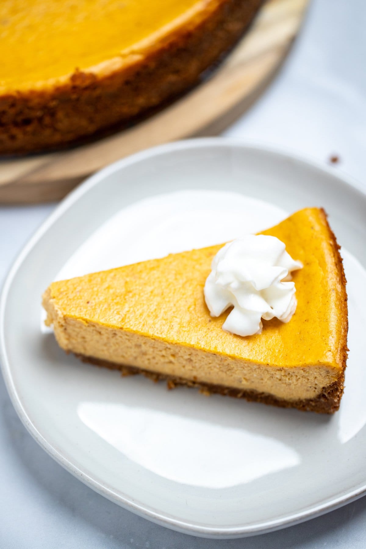 A piece of pumpkin cheesecake on a plate topped with whipped cream, next to the whole cheesecake on a table.