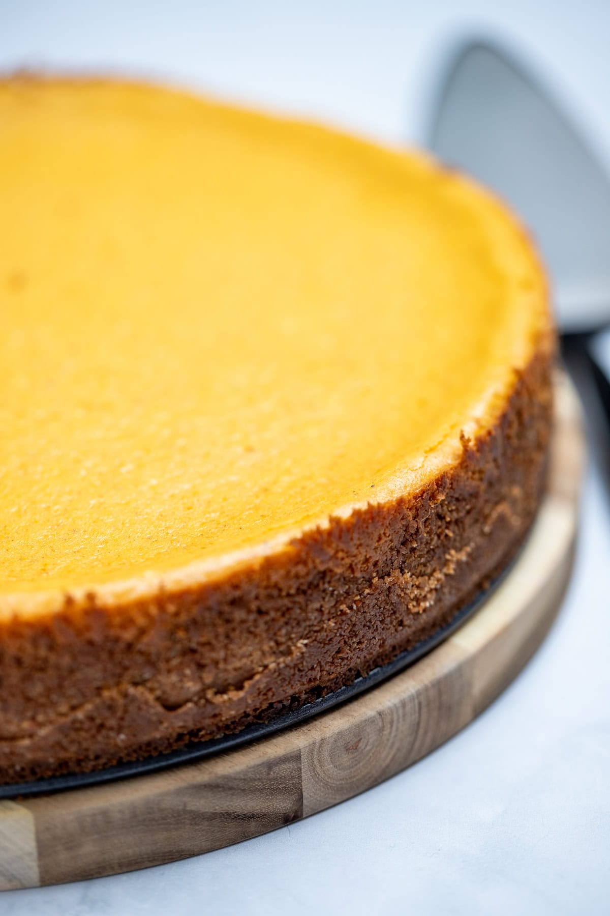 A pumpkin cheesecake sitting on a round wooden cutting board, with a pie server sitting on the table next to it.