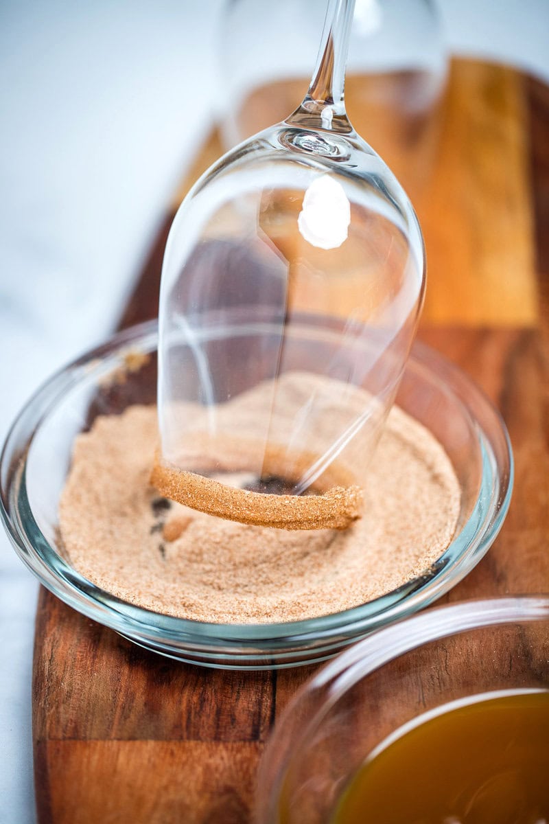 A wine glass with the rim coated in caramel sauce being dipped into a shallow bowl of cinnamon sugar. The bowl is sitting on a cutting board, in front of the shallow bowl of caramel sauce, with another wine glass behind everything.