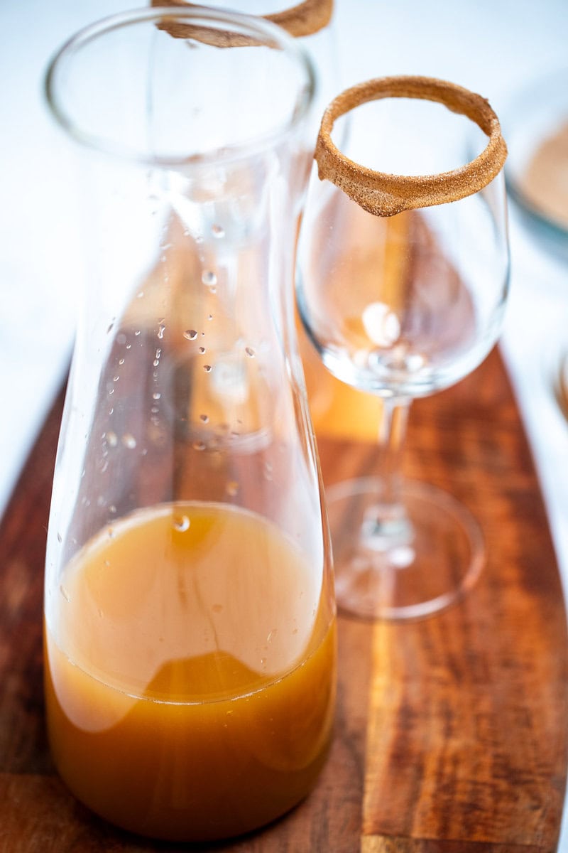 A tall glass container with apple cider filled less than halfway up the container, sitting on a cutting board with wine glasses behind it. The wine glasses have rims coated with caramel and cinnamon sugar.